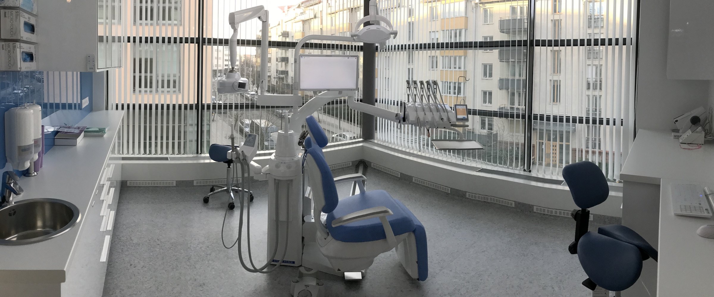 Ljust behandlingsrum med hörnfönster, blå tandläkarstol och handfat på Renaissance Dental