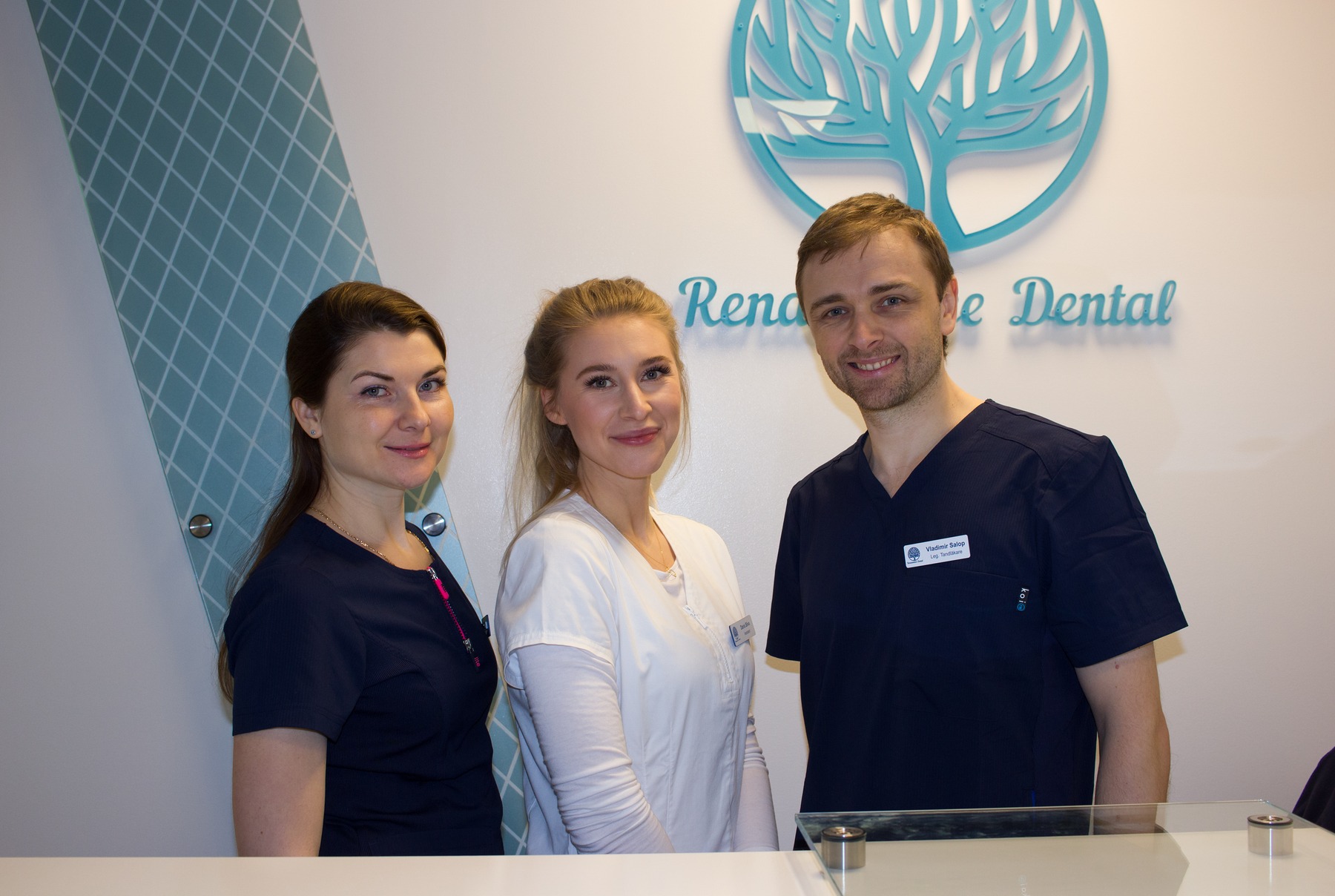 Tre i personalen ler mot kameran bakom receptionsdisken på Renaissance Dental, med klinikens logga på väggen bakom